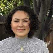 woman with short brown hair against tree. she is wearing a gray sweater and a pendant necklace. 