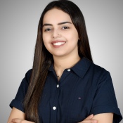 Portrait of a smiling young woman with long, straight hair, wearing a dark blue shirt, standing with her arms crossed against a plain background.